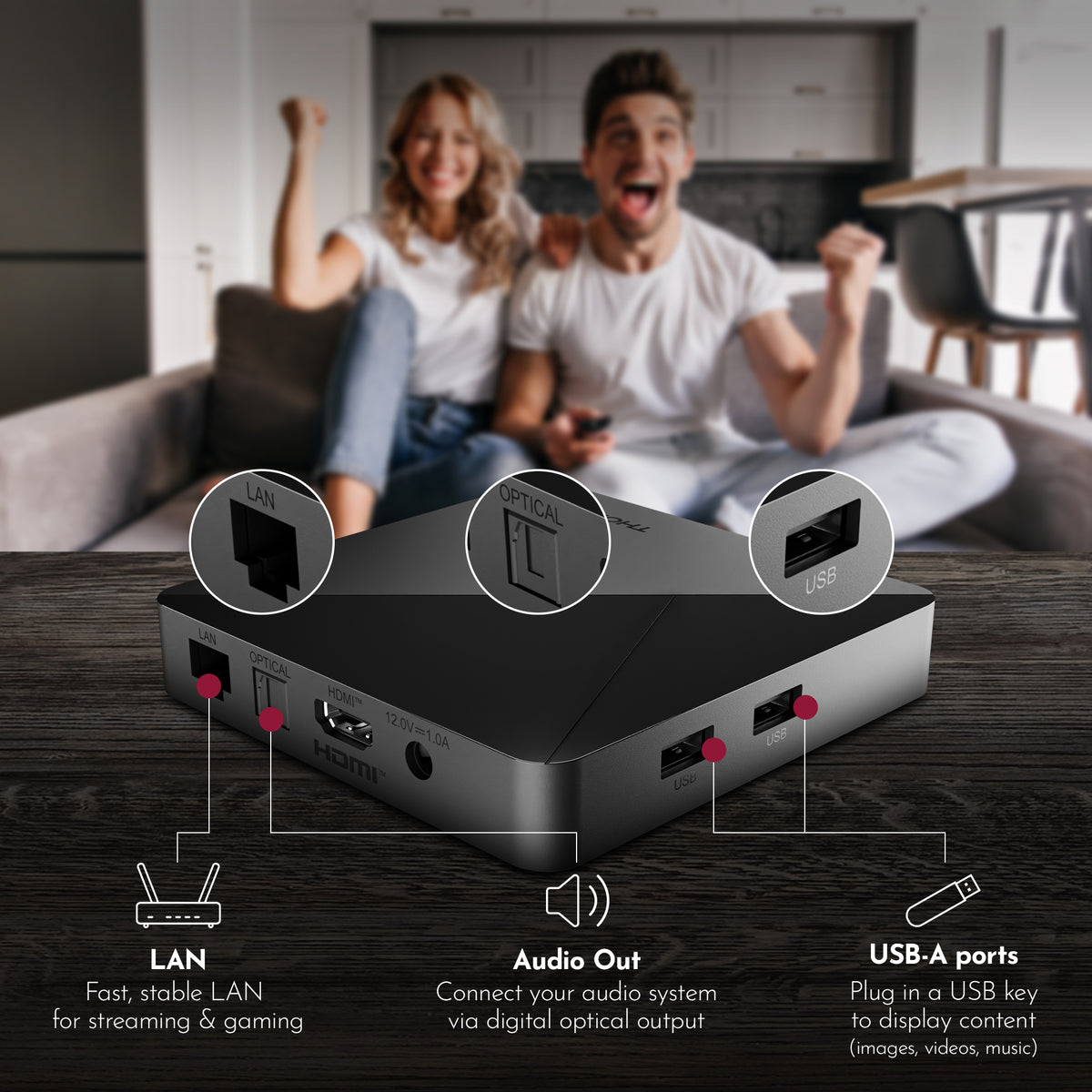 Black streaming device with ports labeled on a wooden surface, with a blurred background of two people sitting on a couch.